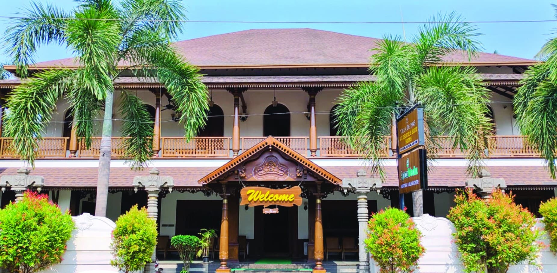 Villa De 1945 by day — a carved timber porch with a Welcome sign, turned timber balustrades on the upper verandah and the Villa De 1945 and AptaVeda signboards